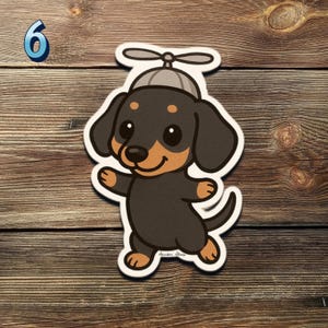 May include: A sticker of a cartoon dachshund dog wearing a propeller hat. The dog is black and tan with a smiling face and outstretched paws. The number 6 is in the upper left corner. The sticker is on a wooden background.