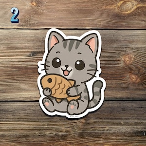 May include: A cute cartoon cat sticker holding a golden-brown fish. The cat is gray with black stripes and big, round eyes. The sticker has a white border and is set against a wooden background. The number "2" is in the top left corner.