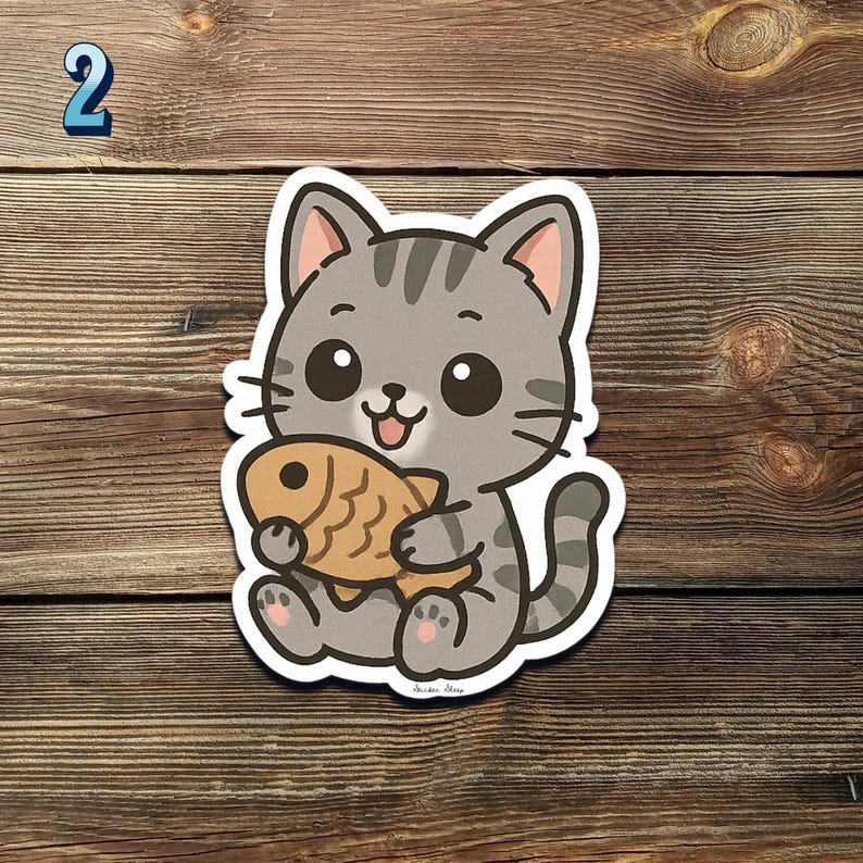 May include: A cute cartoon cat sticker holding a golden-brown fish. The cat is gray with black stripes and big, round eyes. The sticker has a white border and is set against a wooden background. The number "2" is in the top left corner.