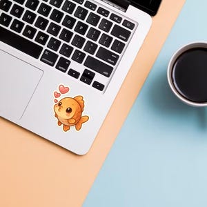 May include: A laptop with a cute orange goldfish sticker blowing kisses, accompanied by two pink hearts. A white mug filled with black coffee sits on a light blue and peach surface.