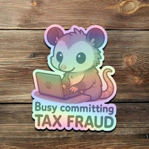 K&ouml;nnte beinhalten: Ein holografischer Aufkleber mit einem Cartoon-Opossum, das einen Laptop benutzt. Das Opossum ist in den Farbt&ouml;nen Lila, Rosa und Gr&uuml;n gehalten. Der Text "Busy committing TAX FRAUD" steht darunter.