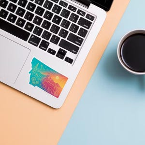 May include: A colorful sticker in the shape of Montana, featuring a bear, mountains, trees, and a sun. The sticker is on a laptop next to a white mug filled with coffee. The background is a mix of peach and light blue.