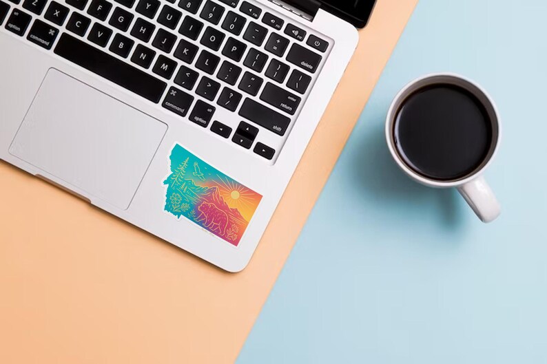 May include: A colorful sticker in the shape of Montana, featuring a bear, mountains, trees, and a sun. The sticker is on a laptop next to a white mug filled with coffee. The background is a mix of peach and light blue.