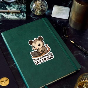 K&ouml;nnte beinhalten: Ein gr&uuml;nes Notizbuch mit einem Aufkleber eines Cartoon-Opossums, das an einem Laptop arbeitet. Der Aufkleber lautet "Busy committing TAX FRAUD". Das Notizbuch liegt auf einer schwarzen Marmoroberfl&auml;che, mit einer Uhr und einem Stift im Hintergrund.