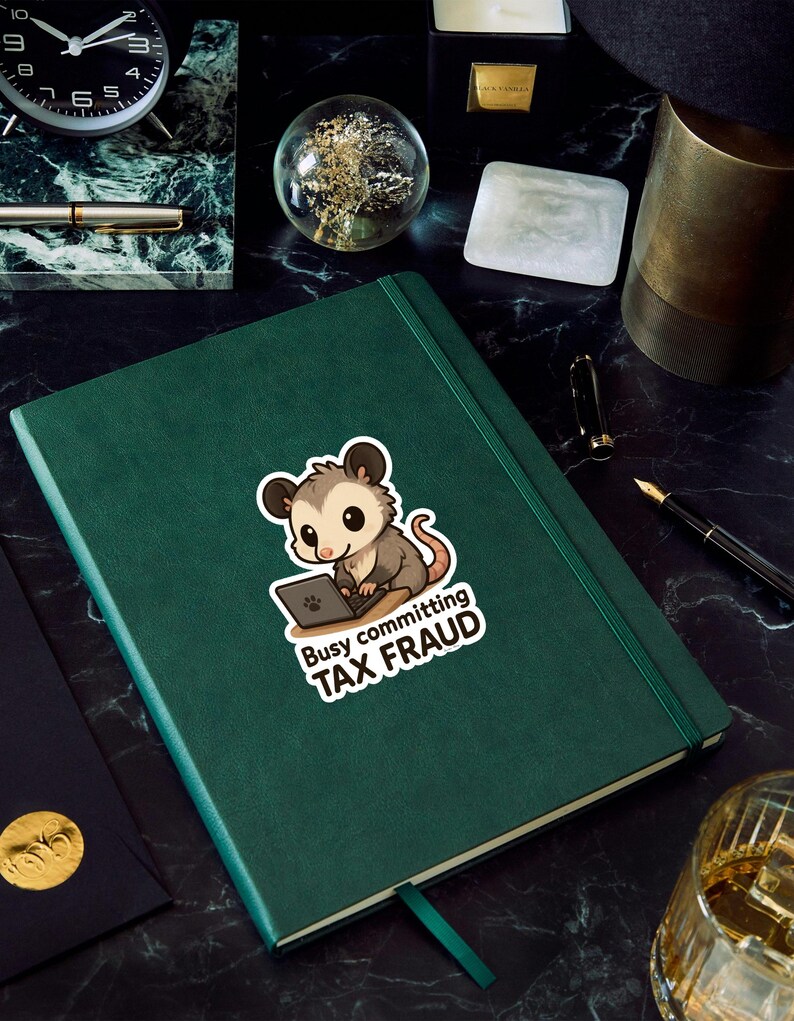 K&ouml;nnte beinhalten: Ein gr&uuml;nes Notizbuch mit einem Aufkleber eines Cartoon-Opossums, das an einem Laptop arbeitet. Der Aufkleber lautet "Busy committing TAX FRAUD". Das Notizbuch liegt auf einer schwarzen Marmoroberfl&auml;che, mit einer Uhr und einem Stift im Hintergrund.