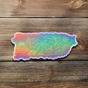 May include: A holographic sticker in the shape of Puerto Rico, featuring a sunrise, lighthouse, palm tree, mountains, a bird, and a turtle. The sticker has a rainbow gradient and is placed on a wooden surface.