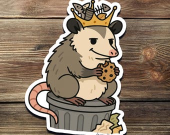 Könnte beinhalten: Ein Cartoon-Opossum, das eine Krone aus Fischknochen trägt und einen Schokoladenkeks hält. Das Opossum sitzt auf einem grauen Mülleimer, aus dem ein brauner Papierbeutel herausragt. Der Text "Sticker Sheep" ist unter dem Mülleimer gedruckt.