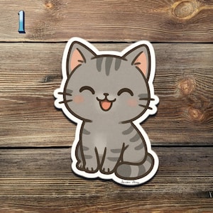 May include: A gray tabby cat sticker with a happy expression. The cartoon cat has pink inner ears and cheeks, with black stripes and whiskers. The sticker is on a wooden surface.