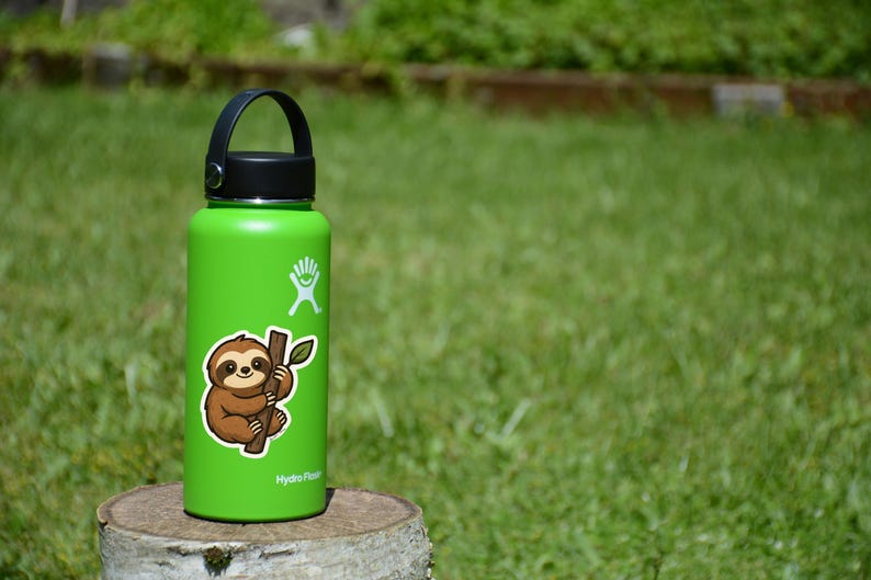 May include: A green insulated water bottle with a black handle and lid. A sticker of a cartoon sloth clinging to a branch is on the bottle. The bottle is sitting on a wooden stump in front of a green grassy background.