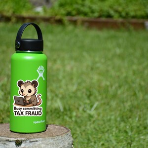 K&ouml;nnte beinhalten: Eine gr&uuml;ne Hydro Flask Wasserflasche mit schwarzem Deckel und Griff. Ein Aufkleber auf der Flasche zeigt ein Cartoon-Opossum mit einem Laptop und dem Text "Busy committing TAX FRAUD."