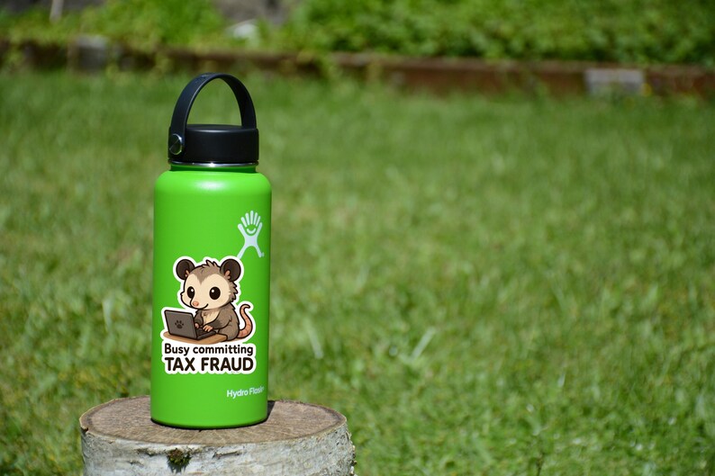 K&ouml;nnte beinhalten: Eine gr&uuml;ne Hydro Flask Wasserflasche mit schwarzem Deckel und Griff. Ein Aufkleber auf der Flasche zeigt ein Cartoon-Opossum mit einem Laptop und dem Text "Busy committing TAX FRAUD."