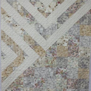 May include: A handmade quilt with a chevron design. The quilt features alternating sections of off-white and patterned fabric in shades of beige, gray, and floral prints. The quilt is machine quilted with a diamond pattern.