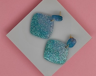 May include: A pair of glittery blue-green earrings. Each earring features a square-shaped main piece and a smaller, rounded accent piece. The earrings are set against a white surface on a pink background.