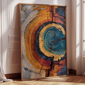 May include: A framed abstract art print featuring a cross-section of a tree trunk. The artwork showcases concentric rings in shades of blue, gold, orange, and red. The print is displayed in a room with a wooden floor and a white wall.