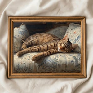 May include: A framed painting of a sleeping tabby cat. The cat is lying on a floral patterned sofa with blue and white accents. The frame is a warm brown color, and the cat has brown and tan stripes.