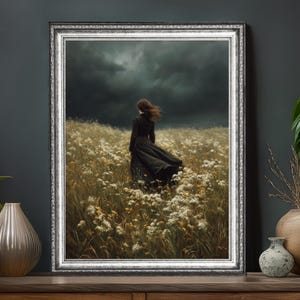 May include: A framed art print depicts a person in a long black dress standing in a field of white and yellow flowers. The artwork has a silver frame and a dark, stormy sky background.