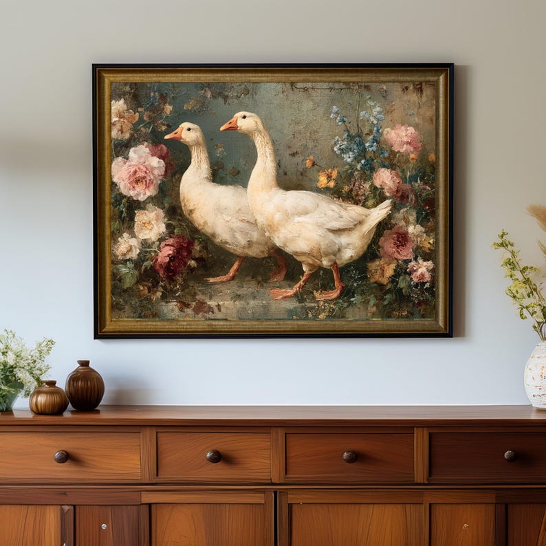 May include: A framed painting depicts two white geese with orange beaks and feet, set against a backdrop of colorful flowers. The artwork features a gold frame and is displayed on a wooden cabinet.