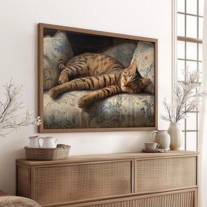 May include: A framed print of a tabby cat lounging on a floral patterned armchair. The cat has brown and black stripes and is lying down with its paws extended. The artwork is displayed above a wooden cabinet with decorative items.