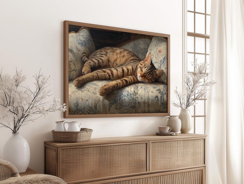 May include: A framed print of a tabby cat lounging on a floral patterned armchair. The cat has brown and black stripes and is lying down with its paws extended. The artwork is displayed above a wooden cabinet with decorative items.