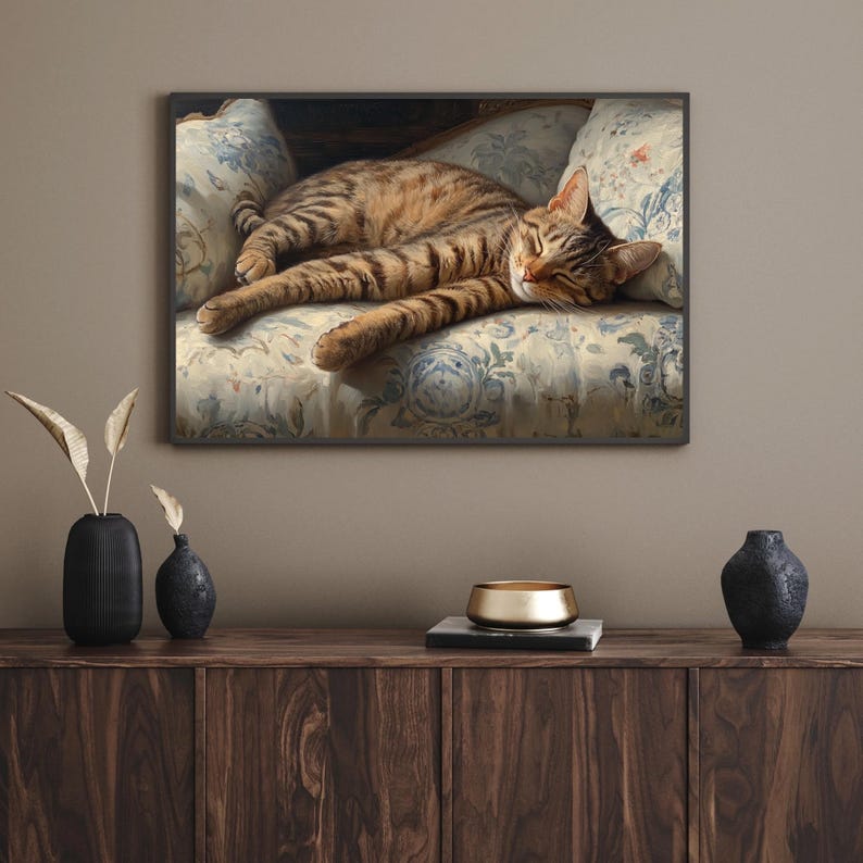 May include: A framed painting of a tabby cat resting on a floral patterned cushion. The cat has brown and black stripes and is lying down with its eyes closed. The cushion has a light blue and white floral design.