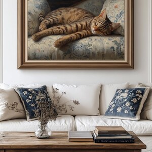 May include: A framed painting depicts a sleeping tabby cat on a floral patterned sofa. The artwork is in a wooden frame and hangs above a white sofa with decorative pillows. A wooden coffee table with books and a vase of dried flowers is in front of the sofa.
