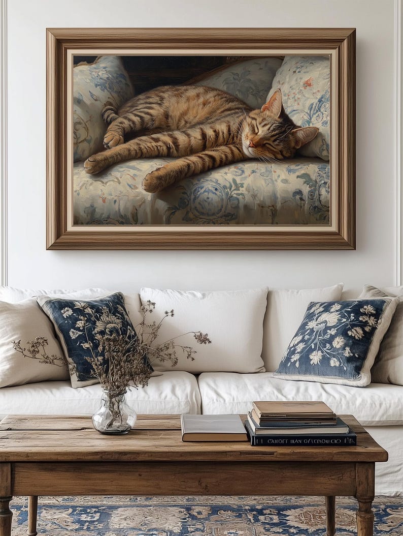 May include: A framed painting depicts a sleeping tabby cat on a floral patterned sofa. The artwork is in a wooden frame and hangs above a white sofa with decorative pillows. A wooden coffee table with books and a vase of dried flowers is in front of the sofa.