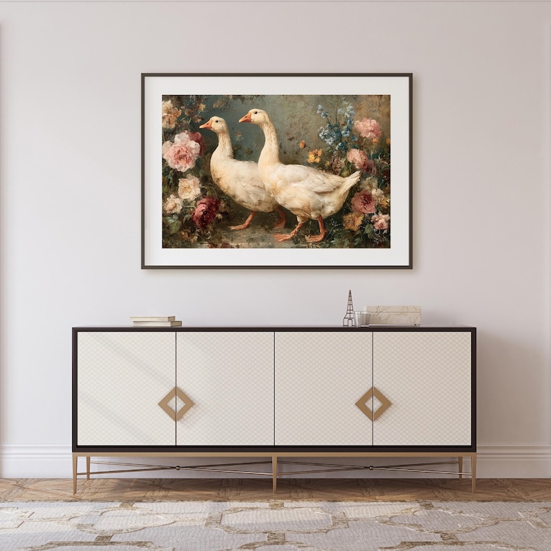 May include: Framed artwork featuring two white geese with orange beaks and feet, set against a backdrop of colorful flowers. The painting is displayed above a white cabinet with gold accents, creating a classic and elegant aesthetic.