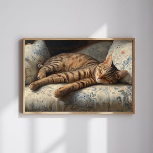 May include: A framed print of a tabby cat sleeping on a floral patterned sofa. The cat has brown and black stripes and is stretched out, appearing relaxed. The sofa cushions have a blue and white floral design, and the frame is a light brown color.