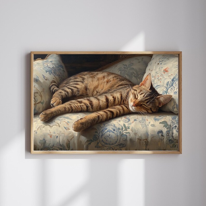 May include: A framed print of a tabby cat sleeping on a floral patterned sofa. The cat has brown and black stripes and is stretched out, appearing relaxed. The sofa cushions have a blue and white floral design, and the frame is a light brown color.