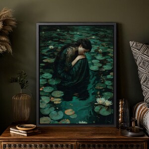 May include: A framed art print depicts a person in a dark green dress, seated in a pond filled with lily pads and white flowers. The artwork has a dark, moody aesthetic, with a focus on the figure's posture and the water's reflections.