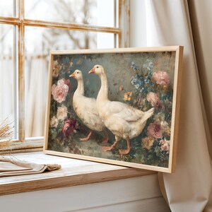 May include: A framed art print featuring two white geese in a garden setting. The artwork has a vintage aesthetic, with soft colors and a floral background of pink, blue, and yellow flowers. The frame is made of light-colored wood.