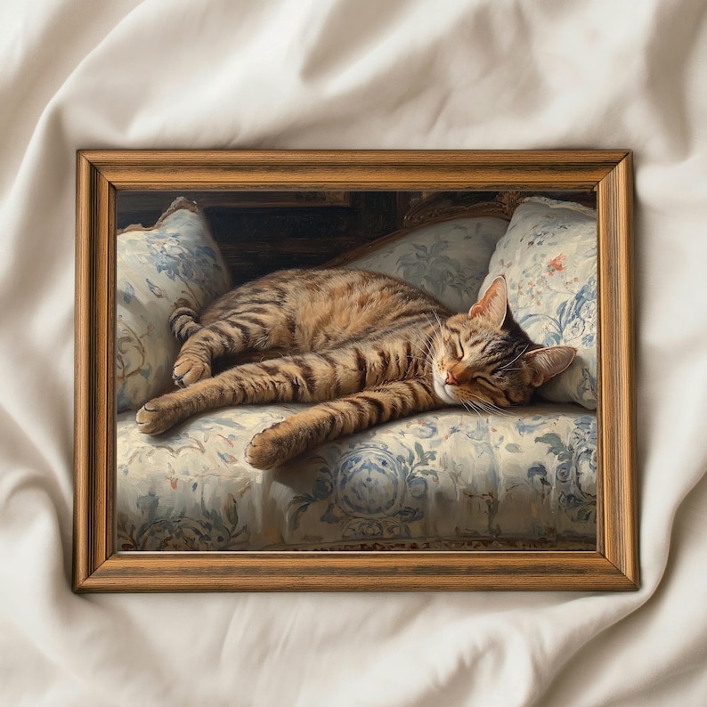 May include: A framed painting of a sleeping tabby cat. The cat is lying on a floral patterned sofa with blue and white accents. The frame is a warm brown color, and the cat has brown and tan stripes.