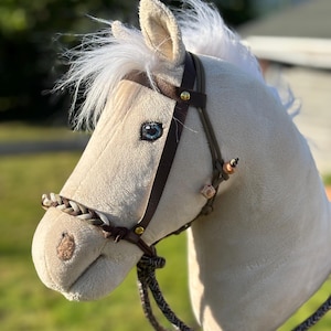 May include: A plush toy horse head in cream with a brown leather bridle and braided reins. The horse has blue eyes, white mane and forelock, and a detailed muzzle. The toy is designed to resemble a real horse.
