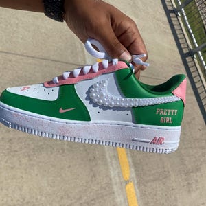 May include: A white, green, and pink sneaker featuring a white pearl-embellished swoosh logo on the side. The shoe includes white laces, a pink heel tab, and the text "PRETTY GIRL" printed in pink on the green side panel above the white sole.