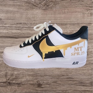 May include: White and black athletic shoe with a gold swoosh and gold dripping paint design. The shoe has the text "MT SPR 25" and "AIR" on the side. The shoe has white laces and a white sole.