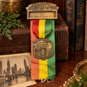 May include: A vintage brass badge featuring a Chicago skyline header with the text CHICAGO, ILL. and REPRESENTATIVE. A vertical ribbon in red, yellow, and green hangs below, attached to a shield-shaped medal with the text 84TH IMPERIAL COUNCIL SESSION, A.A.O.N.M.S., 1958, and IMPERIAL POTENTATE.