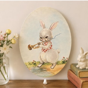 May include: An oval wall hanging featuring a painting of a light gray rabbit wearing round glasses and a red and white checkered vest while playing a gold trumpet. The rabbit stands on a grassy bank near a small stream against a pale blue sky.