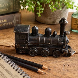 May include: A black metal train figurine with a weathered finish, featuring a tall smokestack, a cab, and multiple wheels. The locomotive has a detailed boiler and a cowcatcher at the front, presented as a solid, stationary decorative object.