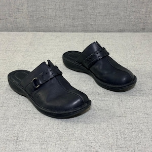 May include: A pair of black leather clogs with a slip-on design. Each clog features a woven strap with a metal buckle and a rounded toe. The shoes have a low heel and a textured sole.