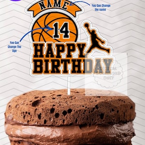 May include: A basketball-themed cake topper with the words "HAPPY BIRTHDAY" in orange and black. The topper includes a basketball graphic, the number 14, and a silhouette of a basketball player. The cake is a layered chocolate cake.