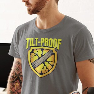 May include: Grey T-shirt featuring the text "TILT-PROOF" above a yellow shield graphic with a cracked design and a grey stripe. A skateboard is held in the person's hand.