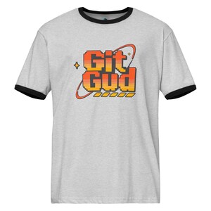 Può includere: T-shirt grigio chiaro con bordi neri su colletto e maniche. La maglietta presenta la scritta "Git Gud" in lettere pixelate arancioni e gialle, con un design retrò e una stella.