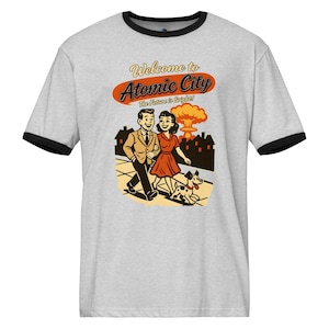 Könnte beinhalten: Hellgraues T-Shirt mit schwarzem Kragen und Bündchen, mit einer Retro-Illustration. Die Grafik zeigt ein Paar, das mit einem Hund spazieren geht, mit einer Atompilzwolke im Hintergrund. Der Text lautet "Welcome to Atomic City" und "The Future is Bright!"