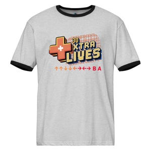 May include: A light gray ringer t-shirt with black trim around the collar and sleeves. The shirt features a retro-style graphic with the text "30 XTRA LIVES" and directional arrows. A plus sign is on the left side of the text.