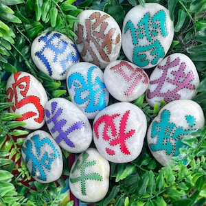 May include: A selection of smooth, white stones, each hand-painted with colourful, stylised symbols. The symbols are outlined with small black dots and feature hues of red, blue, green, purple, and pink. The stones are arranged on a bed of green foliage.