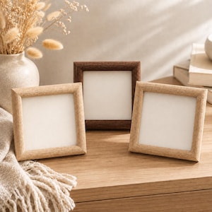 May include: Three square picture frames with wooden borders. Two frames feature a light natural wood finish, while the central frame has a darker brown wood finish. Each frame contains a blank white insert, arranged on a flat wooden surface.