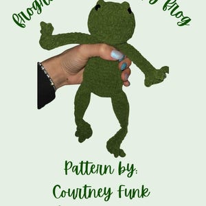 May include: A green crochet frog toy with the text "Frogrick the Grumpy Frog". The frog is held by a hand. The text "Pattern by: Courtney Funk @ life.as.we.craft.it" is also visible. The frog is a handmade item.