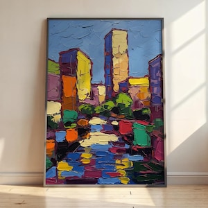 May include: An abstract cityscape painting with bold brushstrokes. The artwork features colorful buildings in shades of yellow, orange, purple, and blue, reflecting in a body of water. The sky is a solid blue, and the painting is framed.