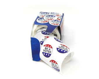 Op de afbeelding: Een rol met blauwe, witte en rode "Be a Voter" stickers in een blauwe en witte doos. De stickers zijn in een cirkel gerangschikt met de tekst naar buiten gericht.