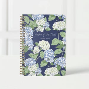 May include: A spiral-bound notebook with a navy blue cover featuring a floral pattern of blue and white hydrangeas and green leaves. The text "Mother of the Bride" and a name are displayed on the cover.